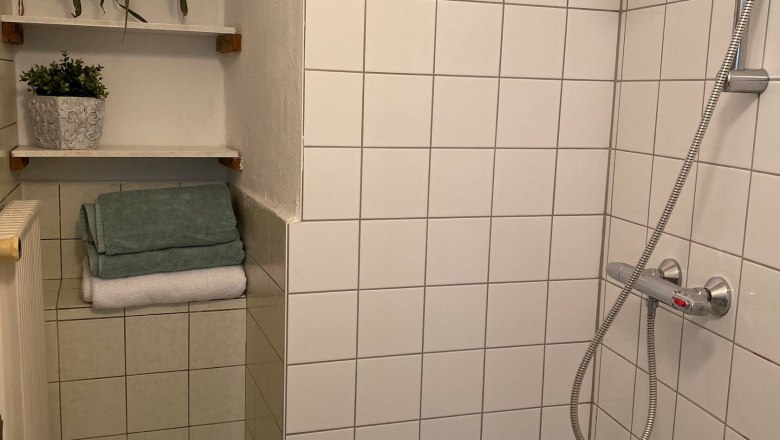 Dusche, © Familie Stagard Eine geflieste Dusche mit Duschkopf, Handtüchern und Pflanzen auf Regalen.