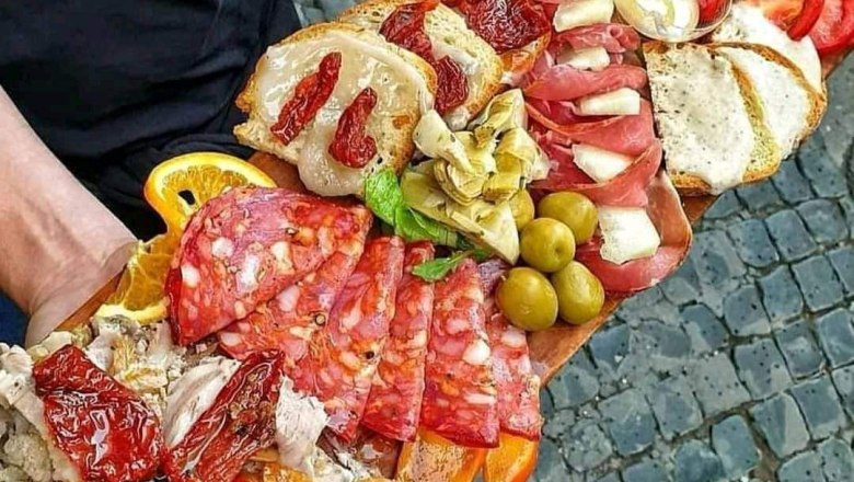Antipasti, © Andy Edlinger Eine große Holzplatte mit verschiedenen Antipasti, darunter Wurst, Käse, Oliven und getrocknete Tomaten, wird von einer Person gehalten.