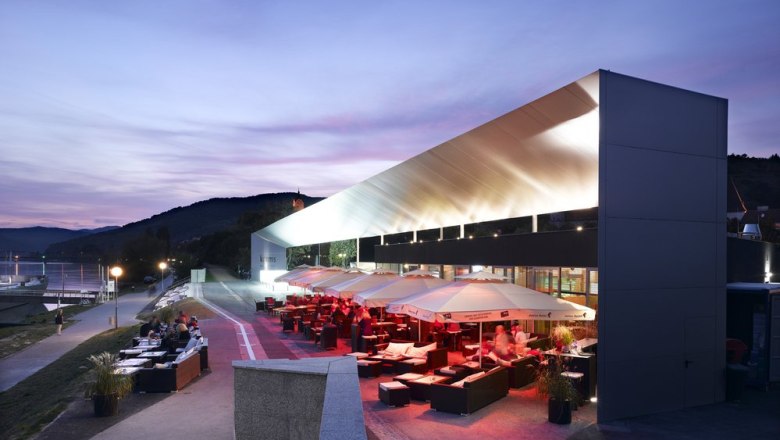 The Wellen.Spiel is located directly on the banks of the Danube., © Wellen.Spiel Modern building on the banks of the Danube at dusk with illuminated terrace and seating areas.