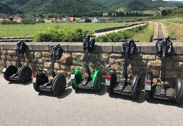 Selection of Segways, © Karoly Six Segways stand in front of a stone wall with a landscape in the background.