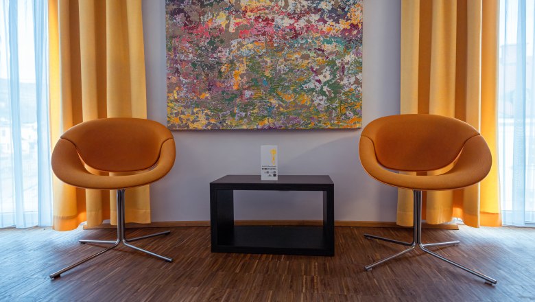 arte Hotel Krems, © Niederösterreich Werbung / Maximilian Pawlikowsky Two orange chairs in front of a colorful painting and yellow curtains.