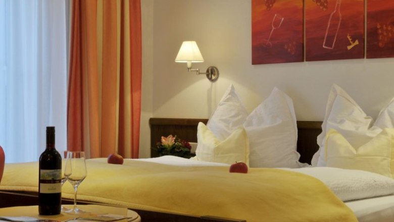 Room example at Weinhof Aufreiter, © Steve Haider Cozy hotel room with double bed, yellow bedspread, wine bottle and glasses on the table, wall art with wine motif.