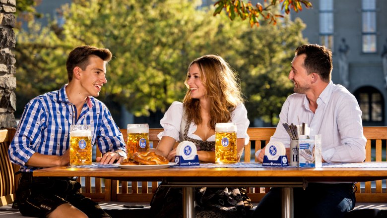 Hofbräu at the Steinertor, © Hofbräu am Steinertor Three people in traditional dress sit at an outdoor table with beer mugs.