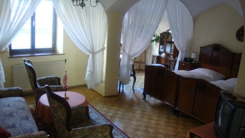 Gästehaus Einzinger GmbH, © Gästehaus Einzinger Ein gemütliches Gästezimmer mit antiken Möbeln, einem Doppelbett, Sesseln und einem kleinen Fernseher. Vorhänge trennen den Schlafbereich ab.