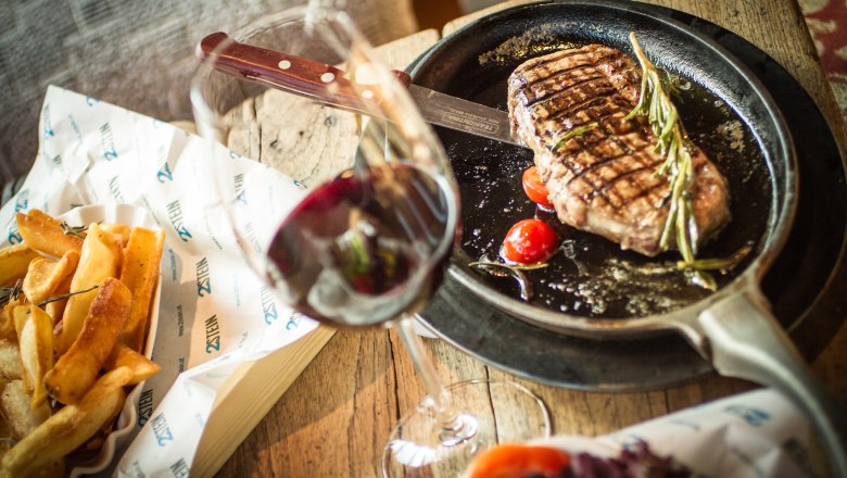 2Stein Bar-Café-Restaurant, © 2stein_Kastner new media Ein Steak in einer Pfanne mit Tomaten und Rosmarin, daneben Pommes und ein Glas Rotwein auf einem Holztisch.