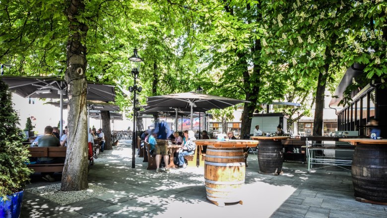 Hofbräu, © Extra Krems Ein Biergarten mit Holztischen und -fässern unter grünen Bäumen, Menschen sitzen und genießen die Atmosphäre.
