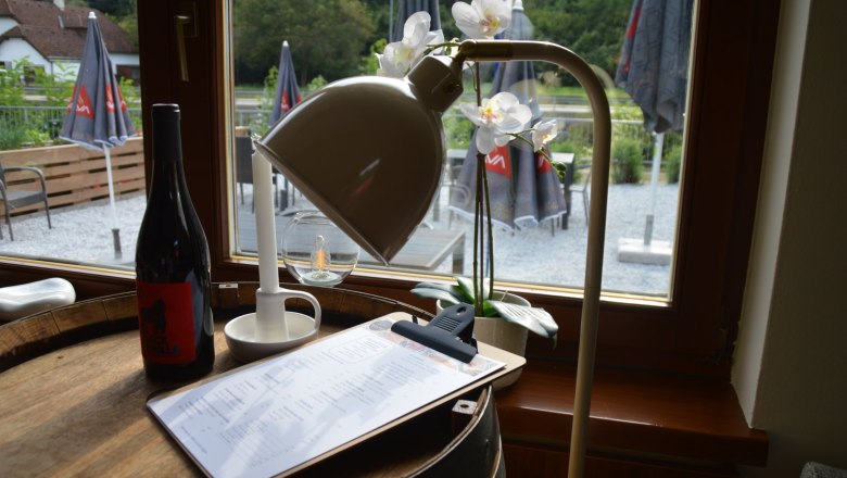 Bistro Rehberg, © Bistro Rehberg Innenansicht eines Bistros mit Weinflasche, Lampe und Menü auf einem Fass, Blick nach draußen auf Terrasse mit Sonnenschirmen.