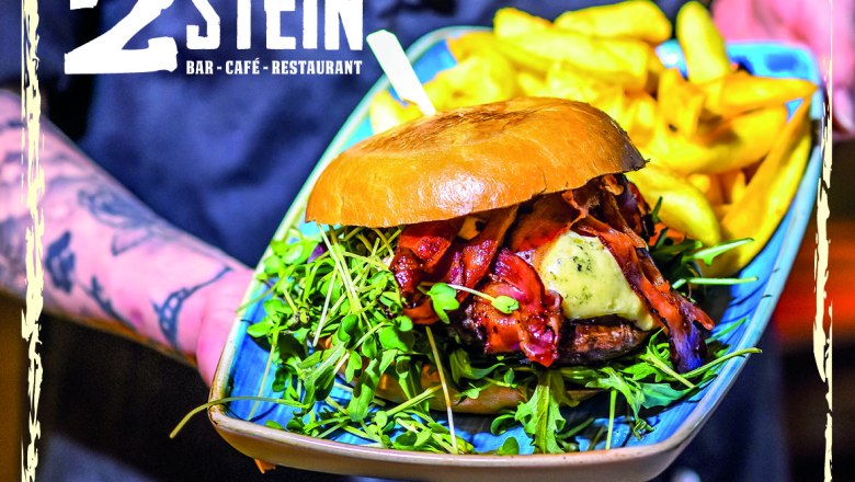 2Stein Bar-Café-Restaurant, © 2Stein Ein Burger mit Speck, Käse und Rucola auf einem Teller mit Pommes, präsentiert von einer tätowierten Hand. Logo von 2Stein Bar-Café-Restaurant oben links.