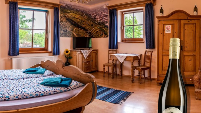 Room no. 5 Barrel bed, © Weingut Zöhrer Cozy room with wooden furniture, a barrel bed and a bottle of wine in the foreground.