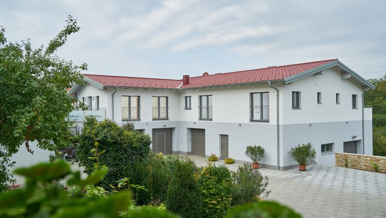 Weinhof Maier, © Weinhof Maier Ein modernes Gebäude mit rotem Dach und weißen Wänden, umgeben von grünen Bäumen und Pflanzen.