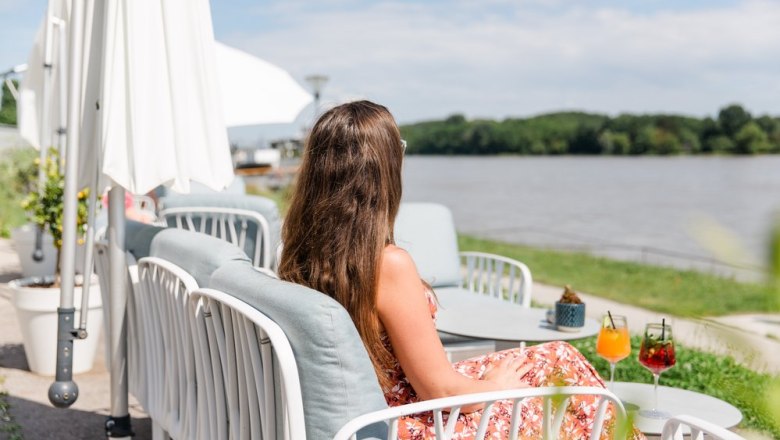 Die Terrasse am Donau-Ufer., © Wellen.Spiel Frau sitzt auf Terrasse am Donau-Ufer mit Getränken auf dem Tisch.