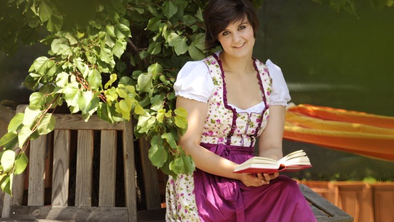 "Zum goldenen Engel" Hotel, Krems, © Haider Woman in traditional dress reading a book on a wooden bench under a tree.