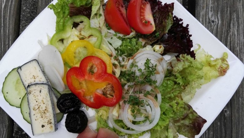 Heurigenjause, © Weinberghof Ilkerl Ein Teller mit Salat, Tomaten, Paprika, Zwiebeln, Schinken und Käse auf einem Holztisch.