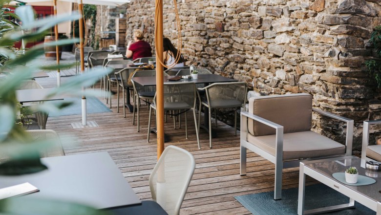 Café living room garden, © FMS Steinertor Gastronomie BetriebsgmbH Cozy outdoor area of a café with wooden floor, modern furniture and stone wall.