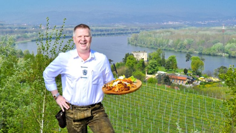 The landlord himself, © Lumpazi Bräu A man in traditional dress holds a tray of food in front of a landscape with a river and vineyards.