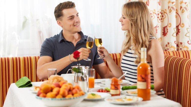 aufreiter_fuehstueck_michael-schafranek, © Michael Schafranek Ein Paar stößt mit Sektgläsern beim Frühstück an, umgeben von Obst und Getränken auf einem gedeckten Tisch.
