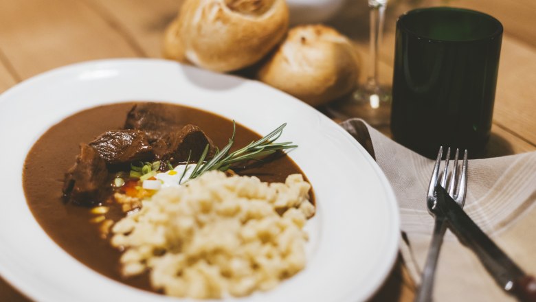 Teller mit Gulasch und Sp&auml;tzle, Brot und Weinglas im Hintergrund.
