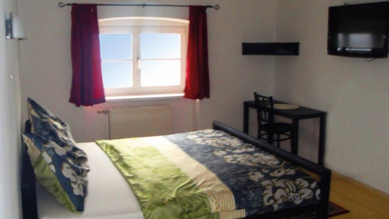 A bedroom with a bed, a window with red curtains, a small table and chair and a TV mounted on the wall.