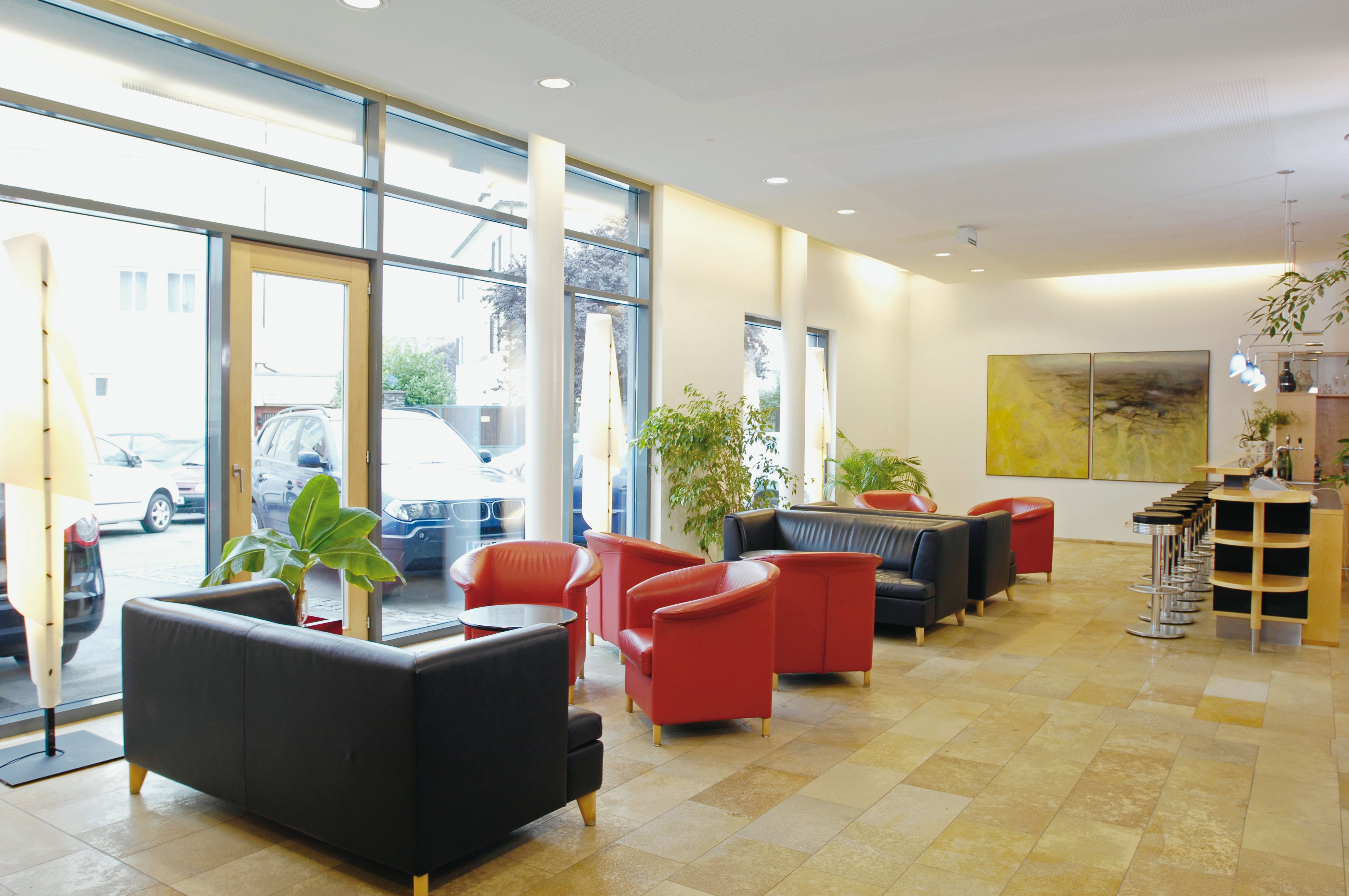 Helle Hotel-Lobby mit roten und schwarzen Sesseln, Barhockern und großen Fenstern.