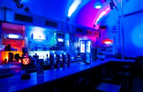 Bar mit blauer und roter Beleuchtung, Flaschen und Gläsern im Regal.