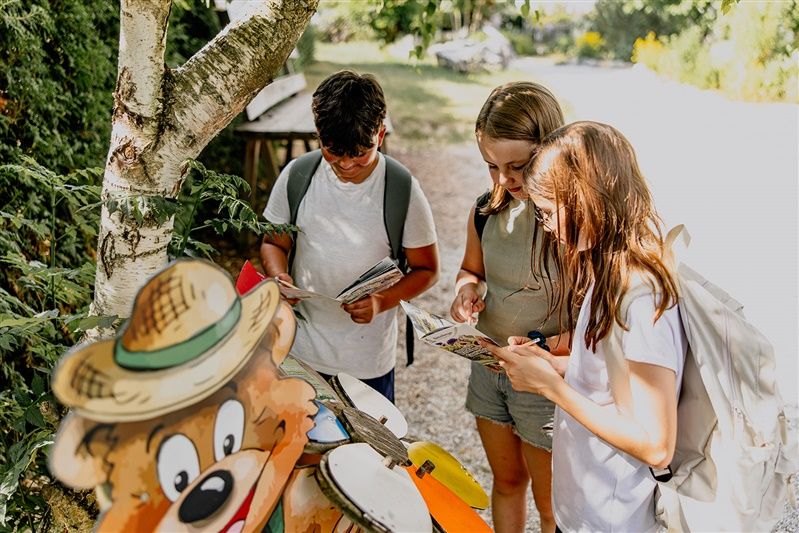 Drei Kinder mit Rucksäcken lesen Karten neben einer Bärenfigur im Freien.