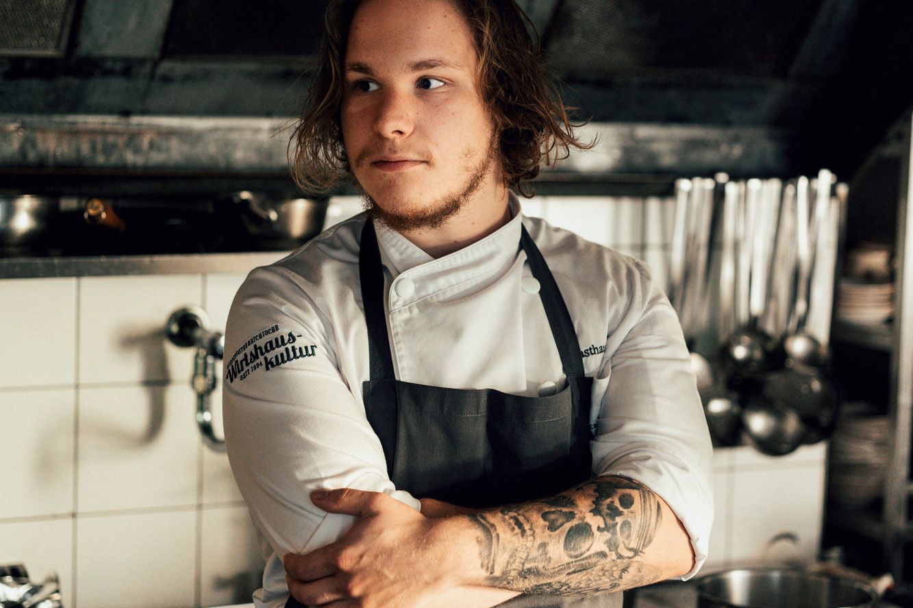 Junger Koch mit Tattoos in einer professionellen Küche, Arme verschränkt.