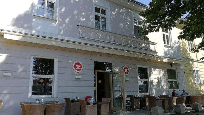 Außenansicht eines Cafés mit Terrasse und Sitzgelegenheiten unter einem Baum.