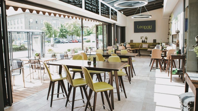 Modern interior of a wine bar with wooden tables and green chairs, large windows and a sign saying 'Leopold'.