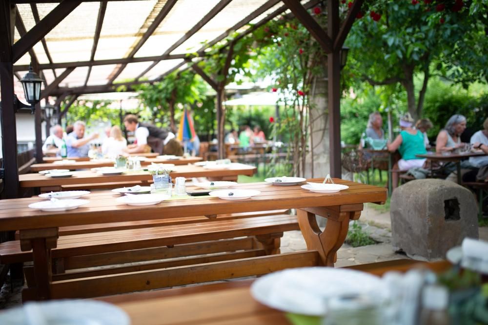 Ein Gastgarten mit Holztischen und -bänken, umgeben von grünen Pflanzen. Menschen sitzen im Hintergrund und genießen ihre Mahlzeiten.