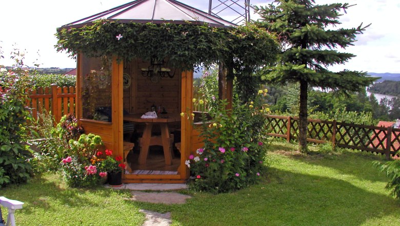 A wooden pavilion in the garden, surrounded by flowers and trees, with a view of a lake in the background.