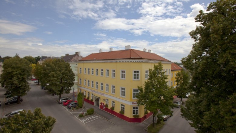 Hotel Unter den Linden Exterior view, © Unter den Linden Exterior view of a yellow hotel with a red roof, surrounded by trees and streets.