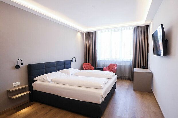 Modernes Hotelzimmer mit Doppelbett, zwei roten Sesseln, einem großen Fenster und einem Flachbildfernseher an der Wand.