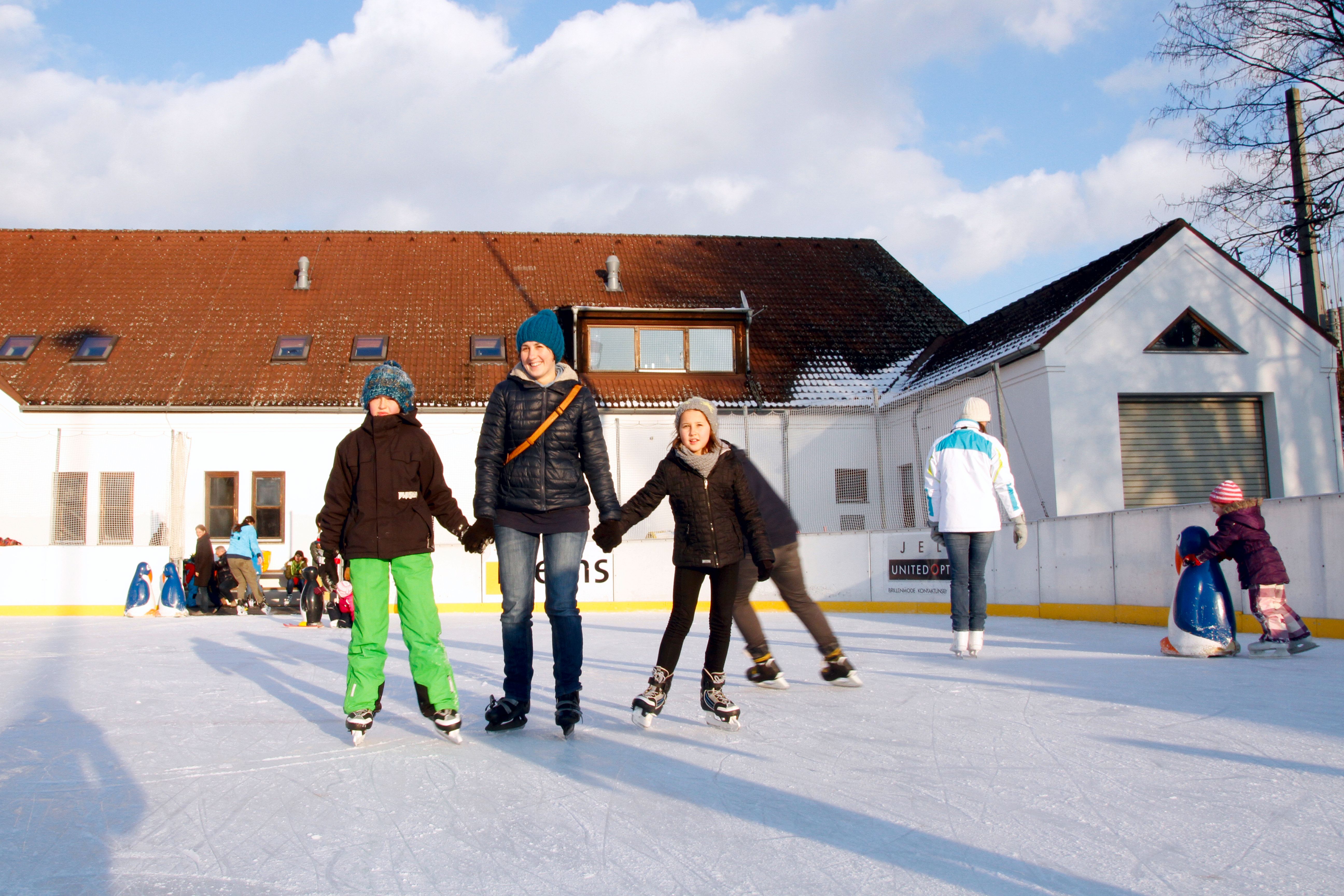Menschen beim Eislaufen auf einer Kunsteislaufbahn vor einem Gebäude.