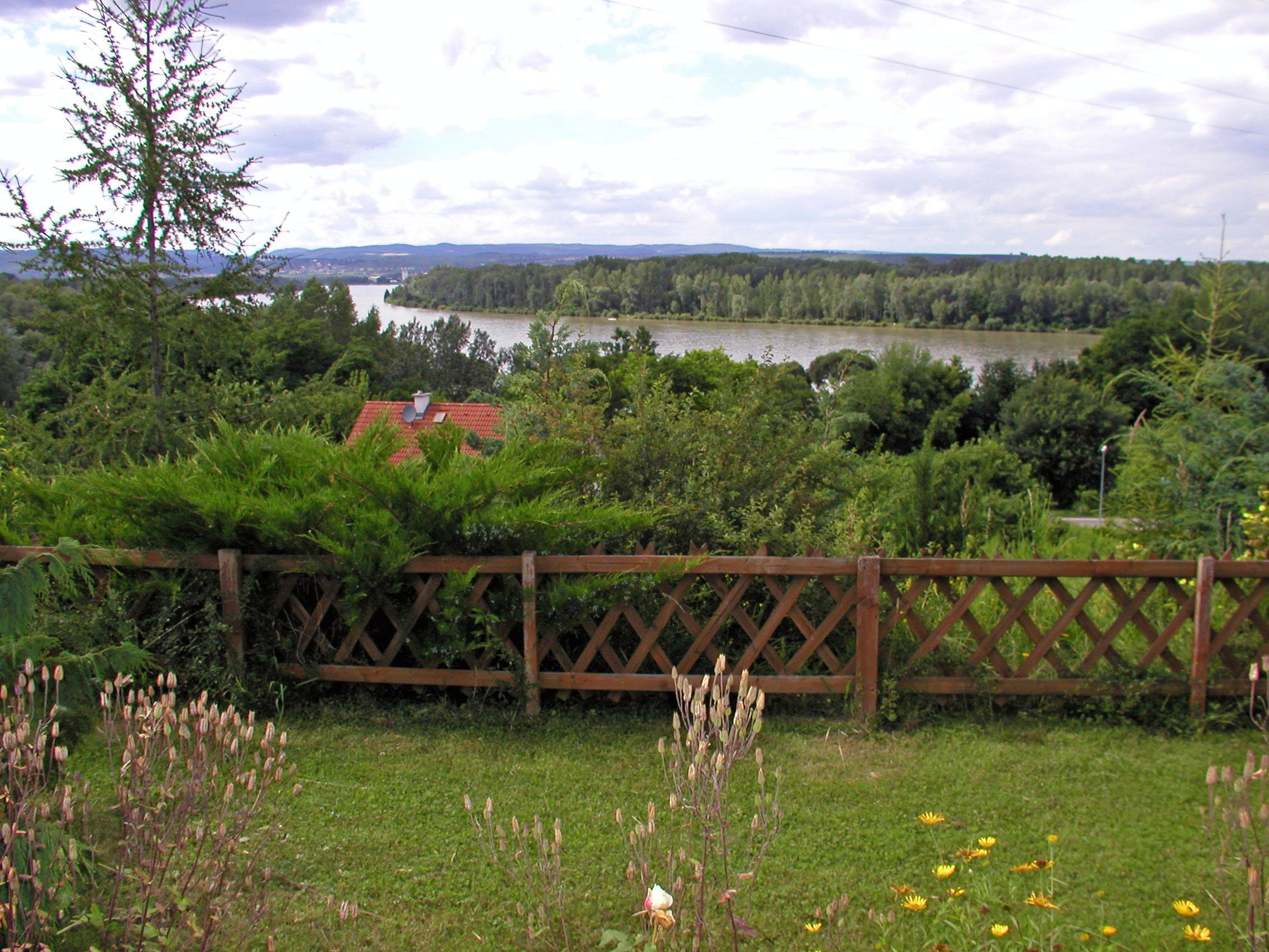 Landschaft mit Fluss, Bäumen und Holzzaun im Vordergrund.