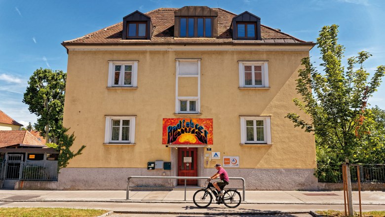 Outside, © Christoph Sammer Yellow building with red entrance and graffiti sign. A cyclist rides past. Blue sky and trees in the background.