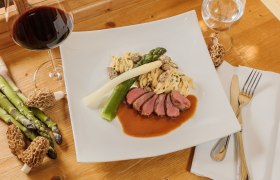 Gourmetgericht mit Fleisch, Spargel, Nudeln und Morcheln auf einem weißen Teller, daneben ein Glas Rotwein.