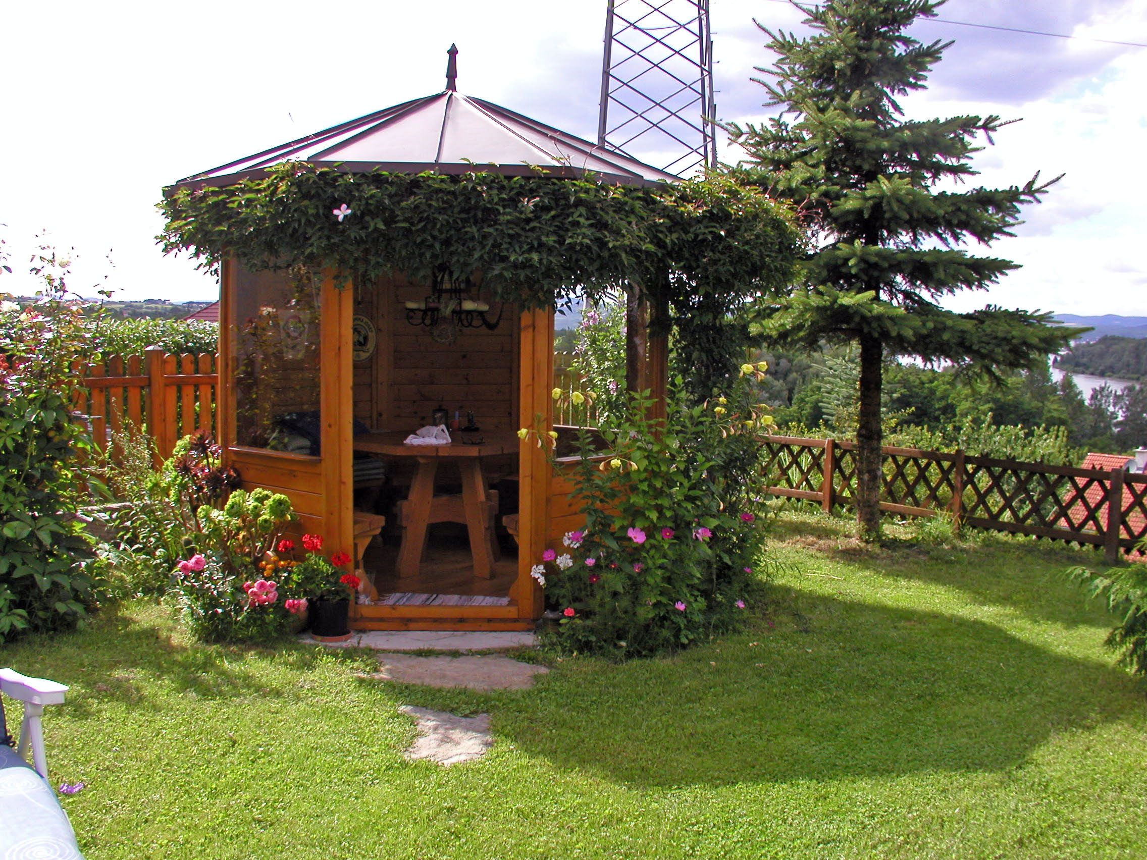 Ein hölzerner Pavillon im Garten, umgeben von Blumen und Bäumen, mit Blick auf einen See im Hintergrund.