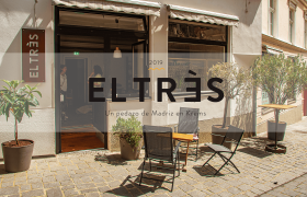 Au&szlig;enansicht eines Caf&eacute;s mit dem Namen EL TR&Egrave;S, Sitzgelegenheiten und Pflanzen vor dem Eingang.
