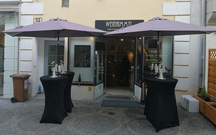 Eingang eines Weinlokals mit zwei Stehtischen und Sonnenschirmen davor.