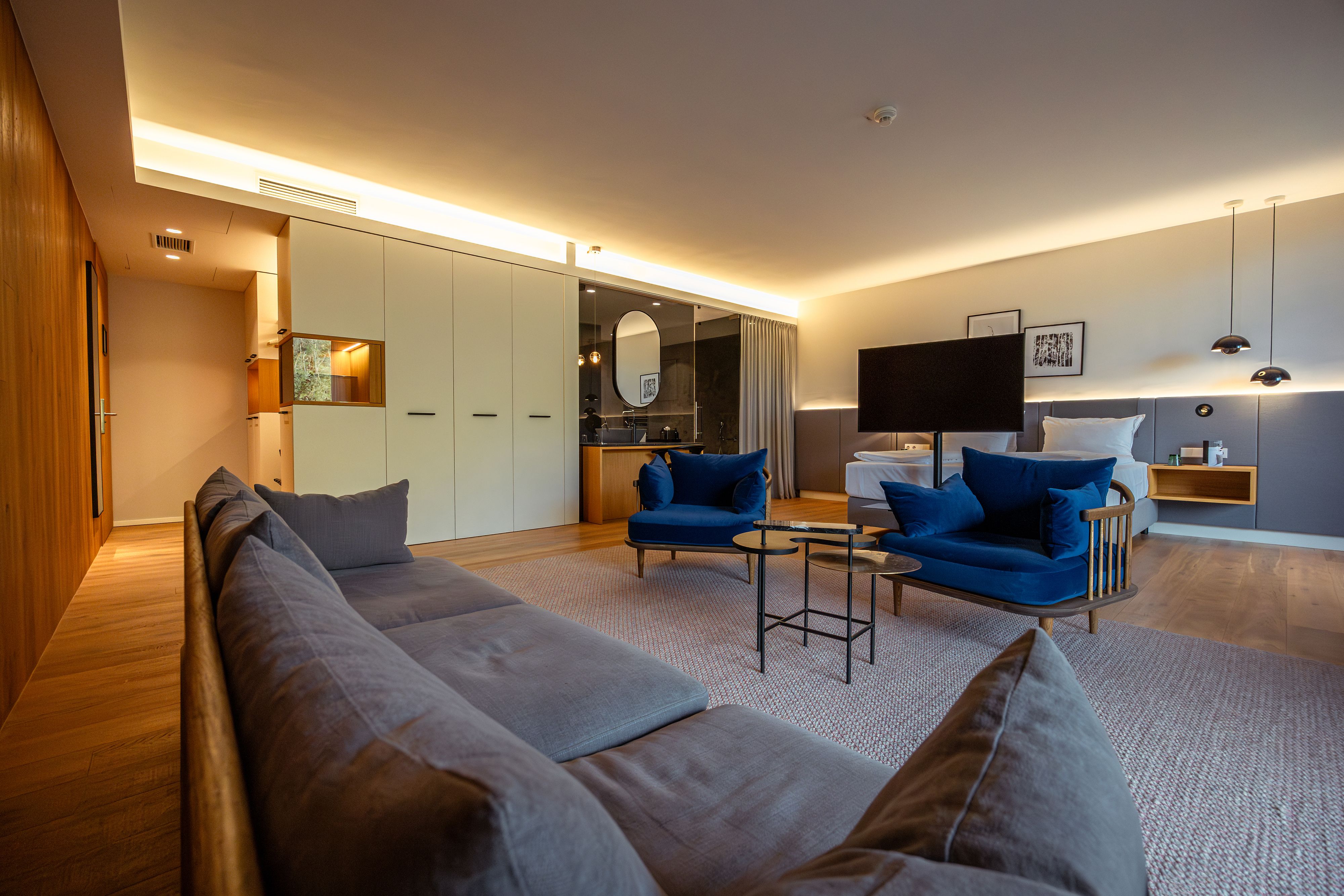 Modernes Hotelzimmer mit blauen Sesseln, grauem Sofa, großem Bett und stilvoller Beleuchtung.