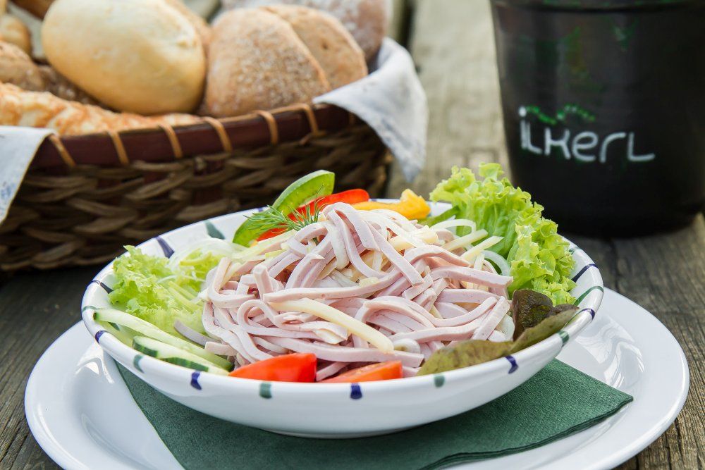Ein Teller mit Wurstsalat, garniert mit Salatblättern, Tomaten und Gurken. Im Hintergrund ein Brotkorb.