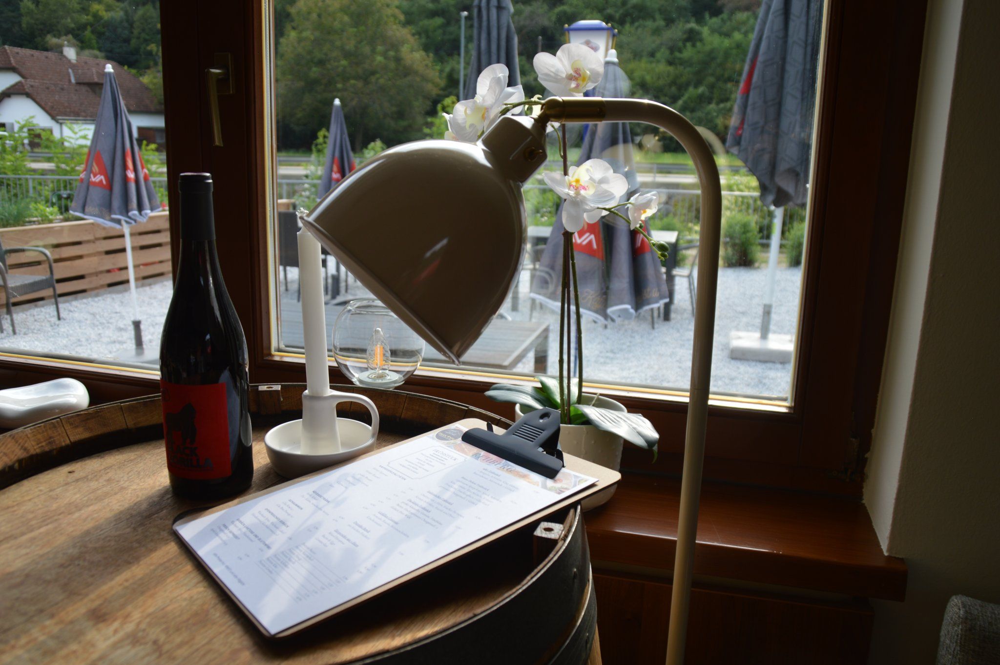 Innenansicht eines Bistros mit Weinflasche, Lampe und Menü auf einem Fass, Blick nach draußen auf Terrasse mit Sonnenschirmen.