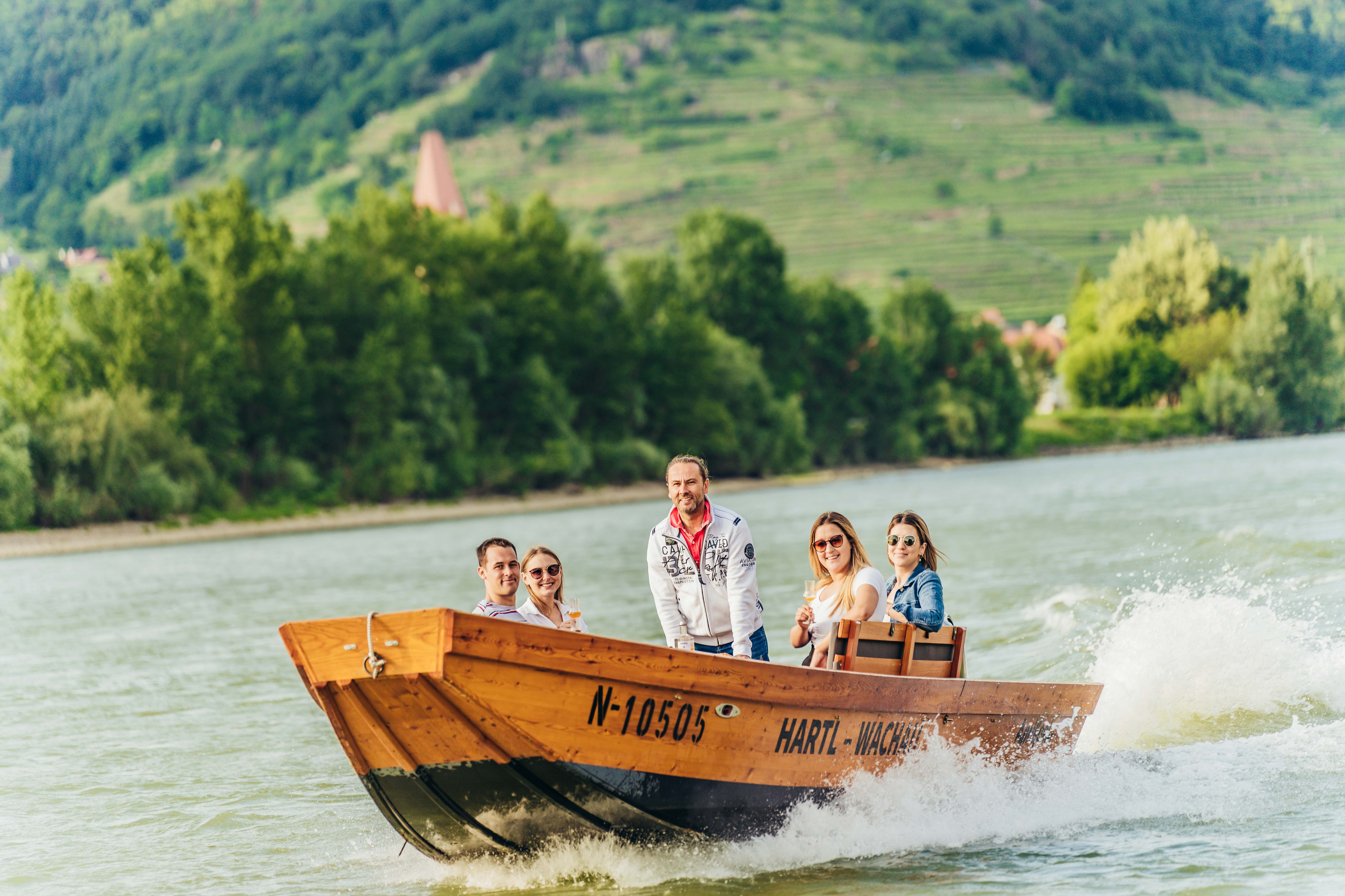 Gruppe von Menschen auf einem Holzboot auf der Donau mit grüner Landschaft im Hintergrund.
