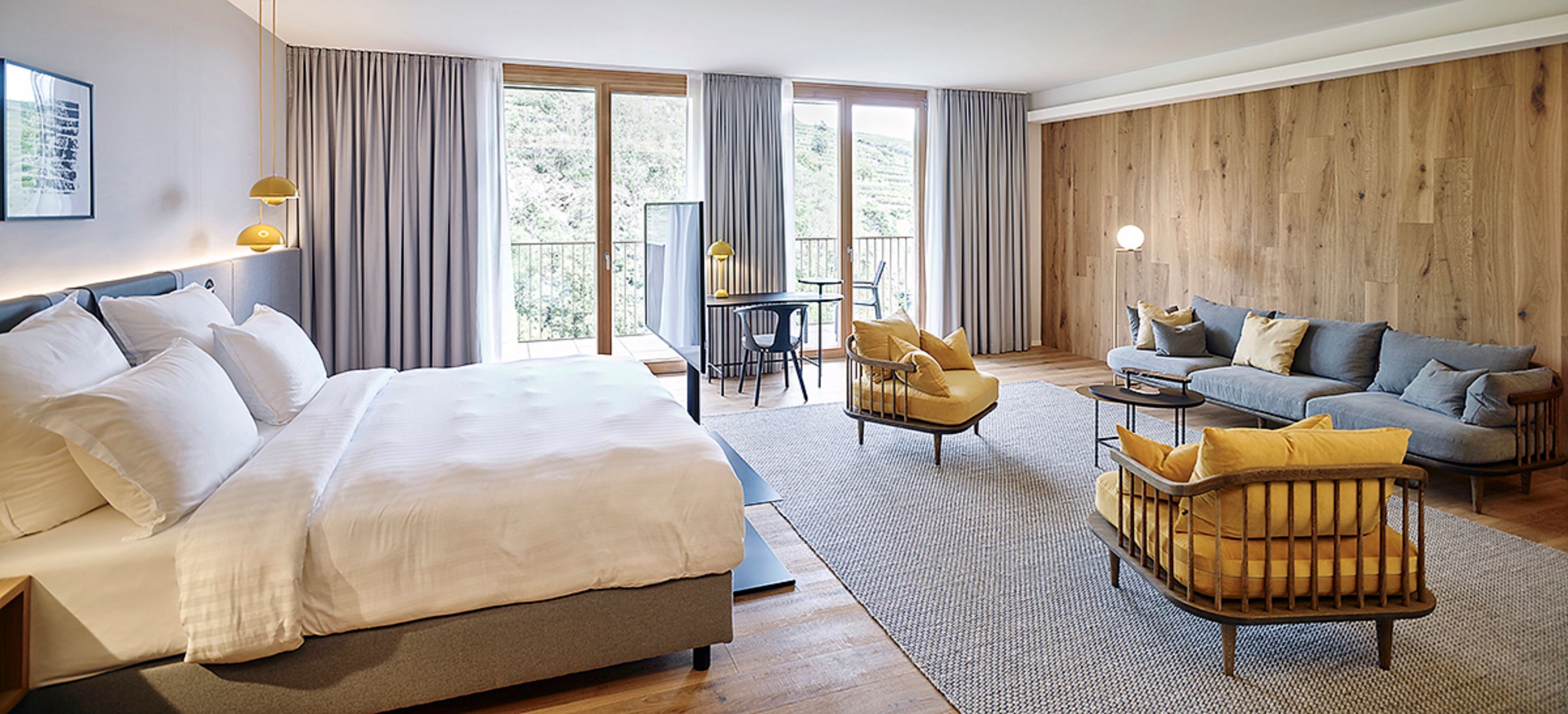 Modernes Hotelzimmer mit großem Bett, Sitzbereich und Schreibtisch vor großen Fenstern.