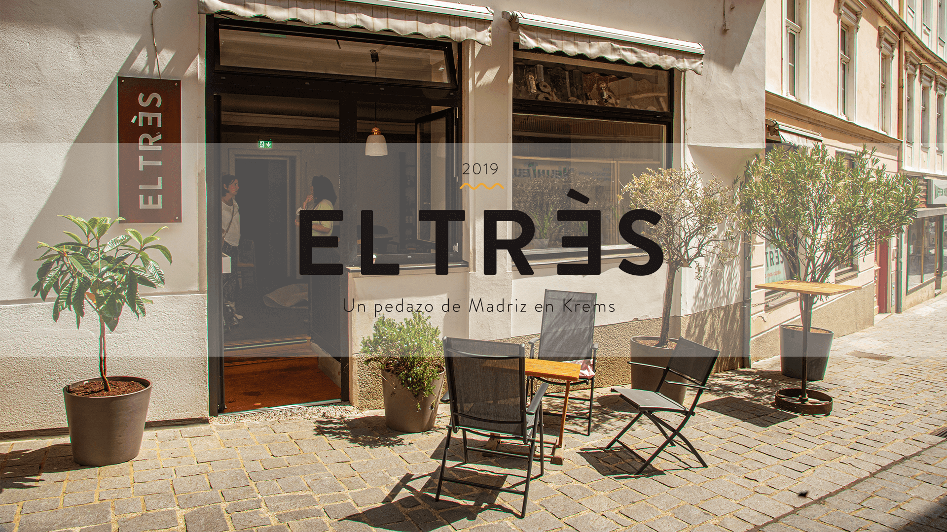 Außenansicht eines Cafés mit dem Namen EL TRÈS, Sitzgelegenheiten und Pflanzen vor dem Eingang.