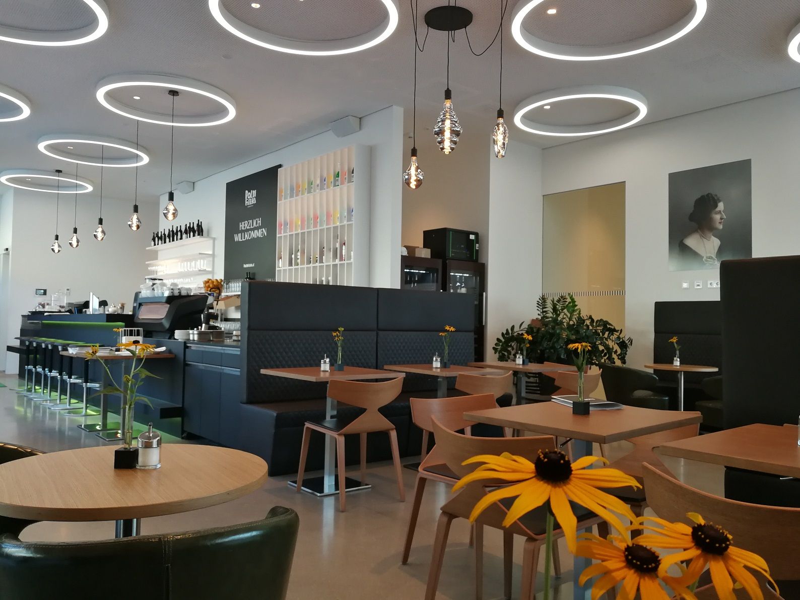 Innenansicht eines modernen Cafés mit runden Deckenleuchten, Holztischen und Blumen auf den Tischen.