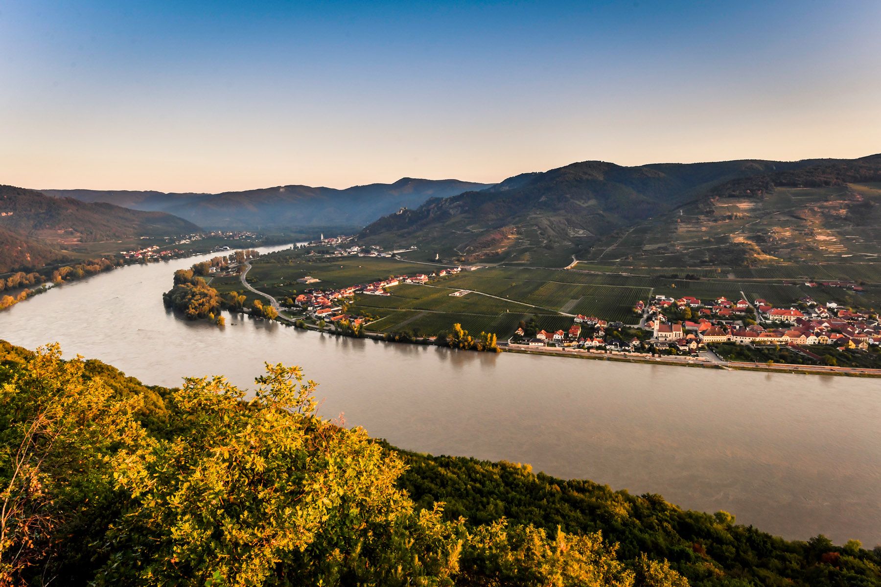 Panoramablick auf einen Fluss, umgeben von Hügeln und Dörfern, bei Sonnenuntergang.