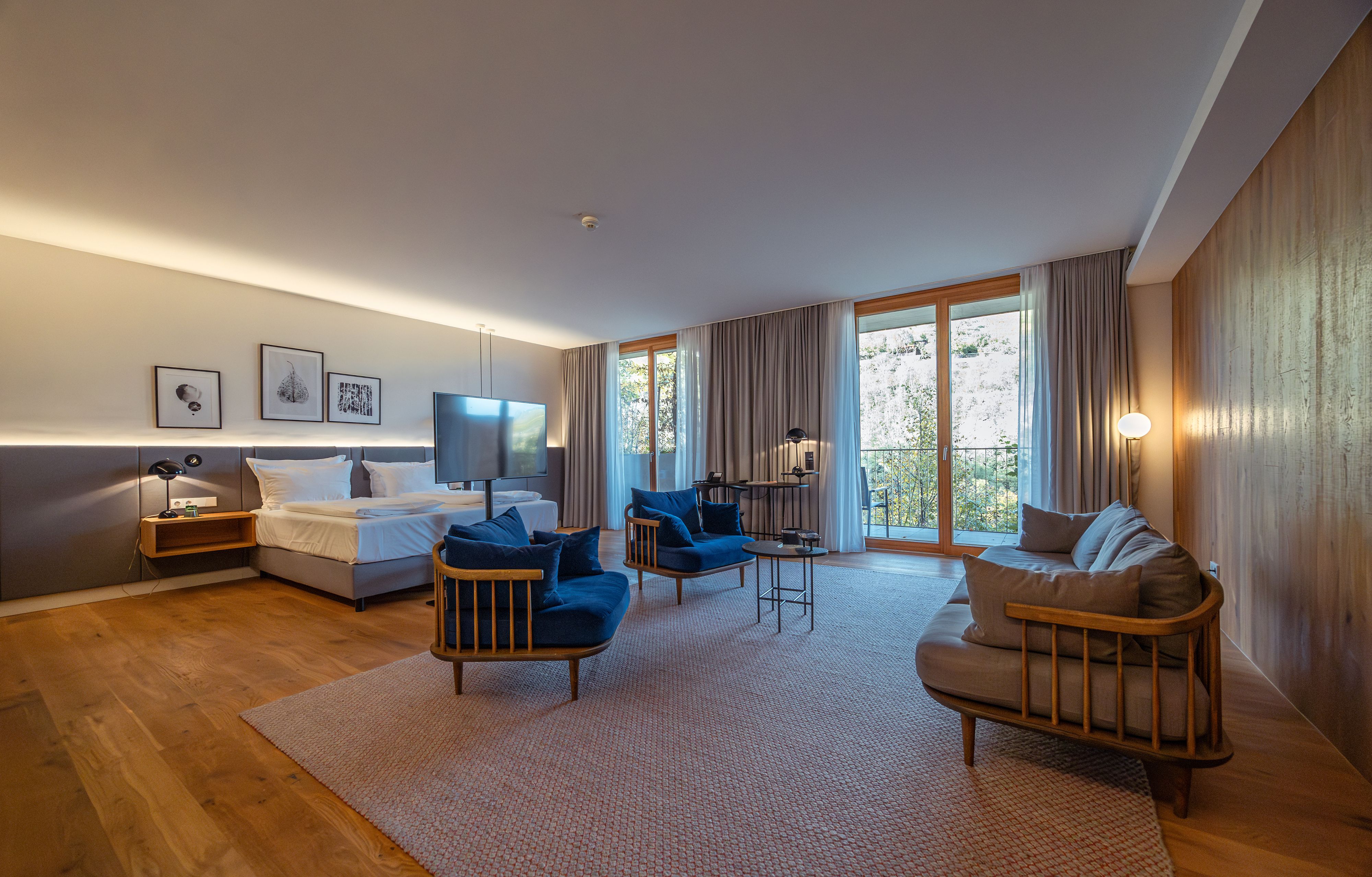 Modernes Hotelzimmer mit Holzboden, großem Bett, blauen Sesseln und großen Fenstern mit Vorhängen.