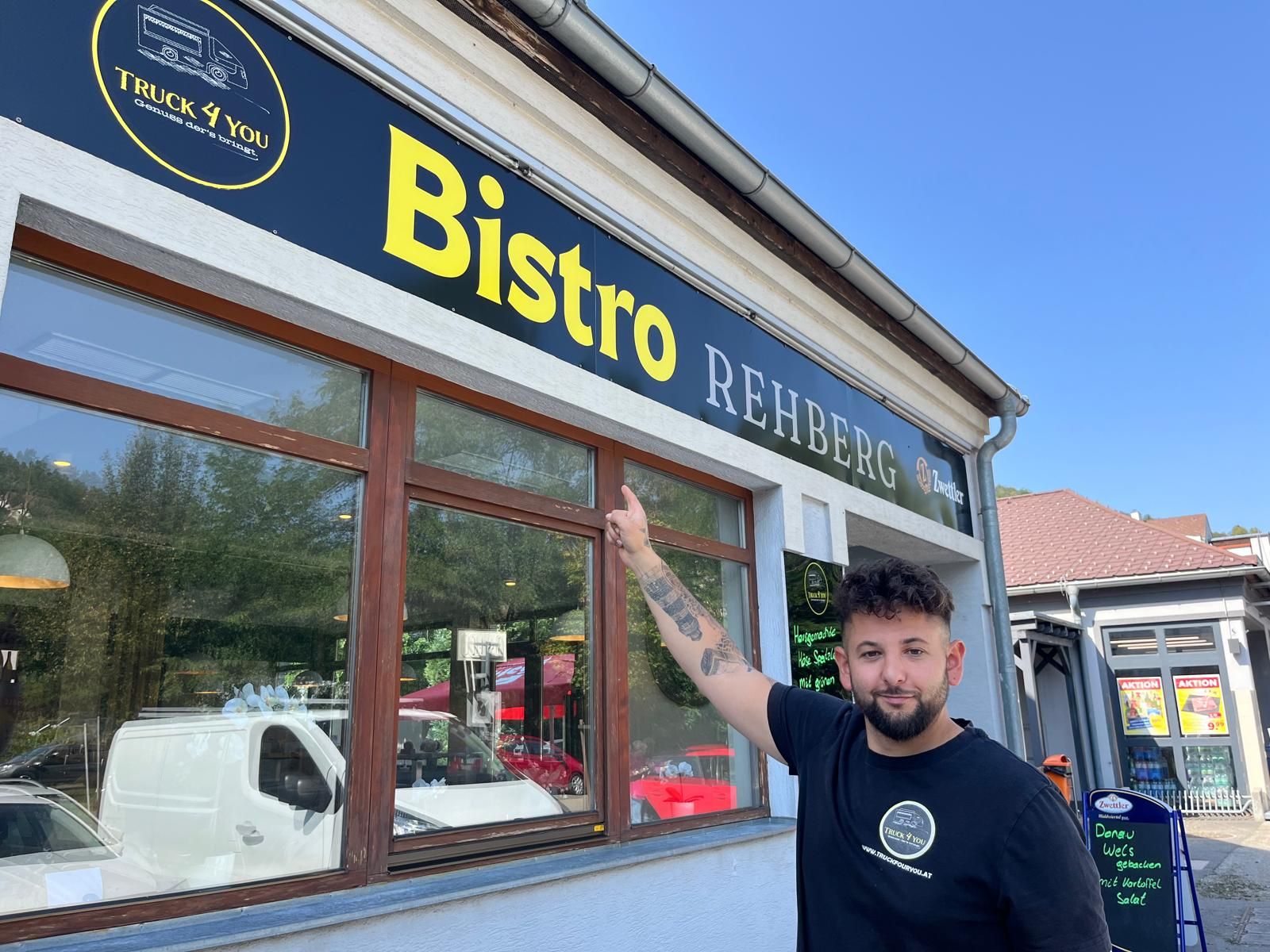 Ein Mann zeigt auf ein Schild mit der Aufschrift 'Bistro Rehberg'.