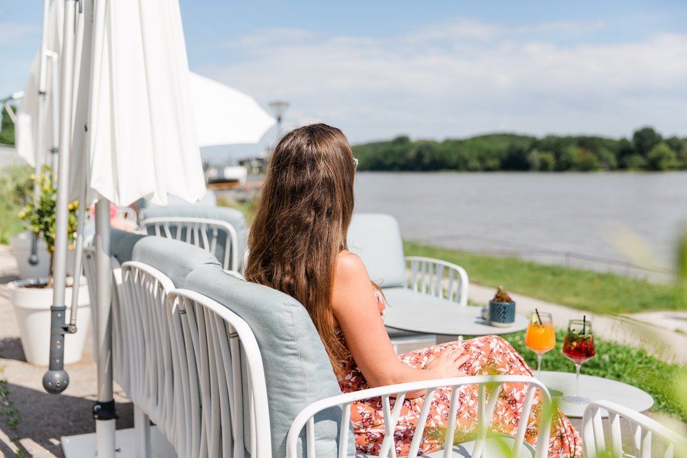 Frau sitzt auf Terrasse am Donau-Ufer mit Getränken auf dem Tisch.